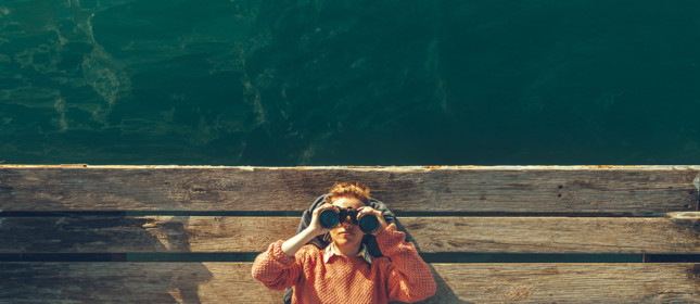 Young Girl In Orange Jacket Lies On A Pier Near The Sea And Looks Through Binoculars. Travel Search Journey Concept